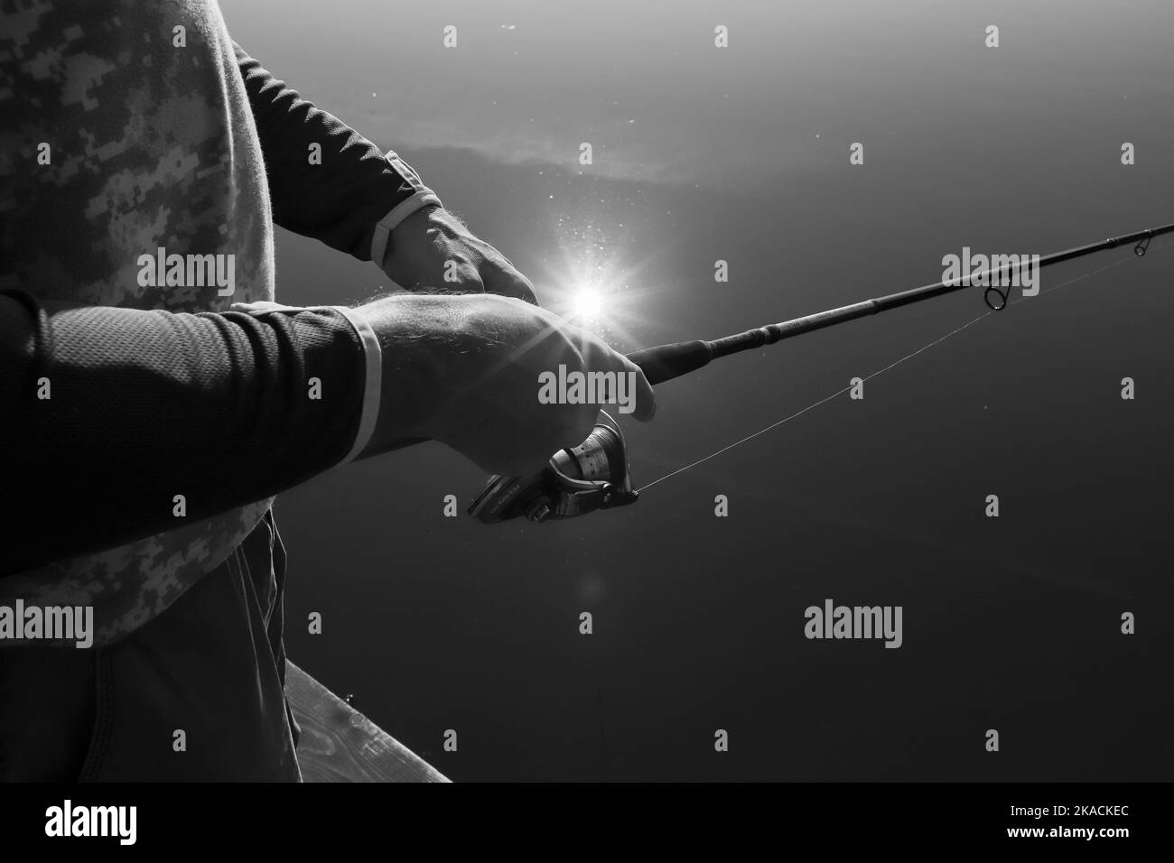 Hands holding fishing rod close up over lake with reflection of sun rays, black and white photo. Stock Photo