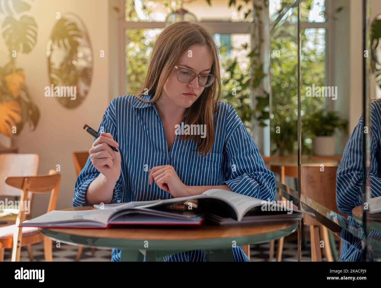 Student in eyeglasses in modern cafe with books and syllabus, studying in trendy coffee shop with day light. Stock Photo