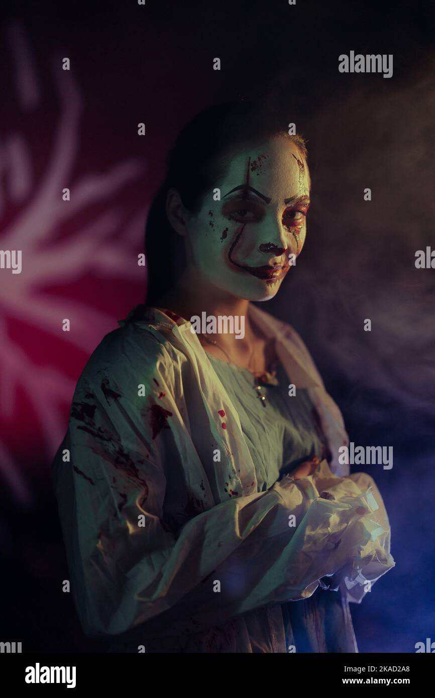 Young woman portrays bloodthirsty zombie with horror wounds on her face and bloody clothes against dark background with lit. Scary image for Halloween Stock Photo