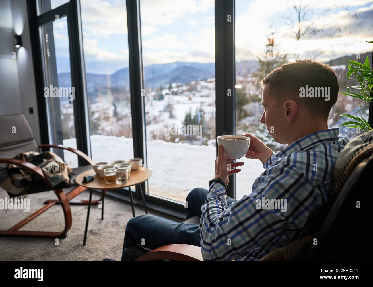 Young man enjoying weekends inside contemporary barn house. Side view of tourist sitting on chair, holding cup of tea, enjoying view of mountain landscape through panoramic windows in new cottage. Stock Photo