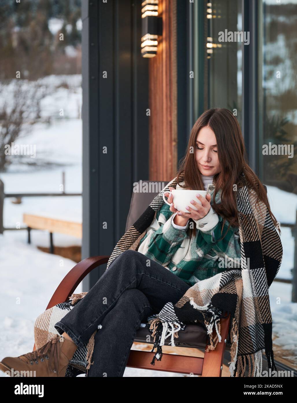 Young woman resting on terrace of modern barn house in the mountains. Happy female tourist sitting in chair, holding cup of tea, enjoying in new cottage in winter. Stock Photo