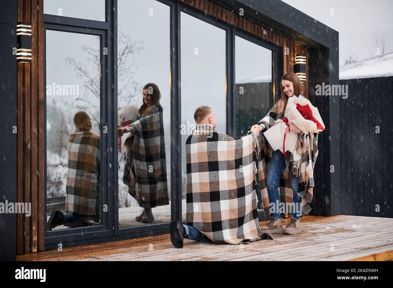 Happy woman holding bouquet of flowers and smiling while man making marriage proposal under winter snow. Couple sharing romantic moment outside scandinavian wooden house barnhouse. Stock Photo