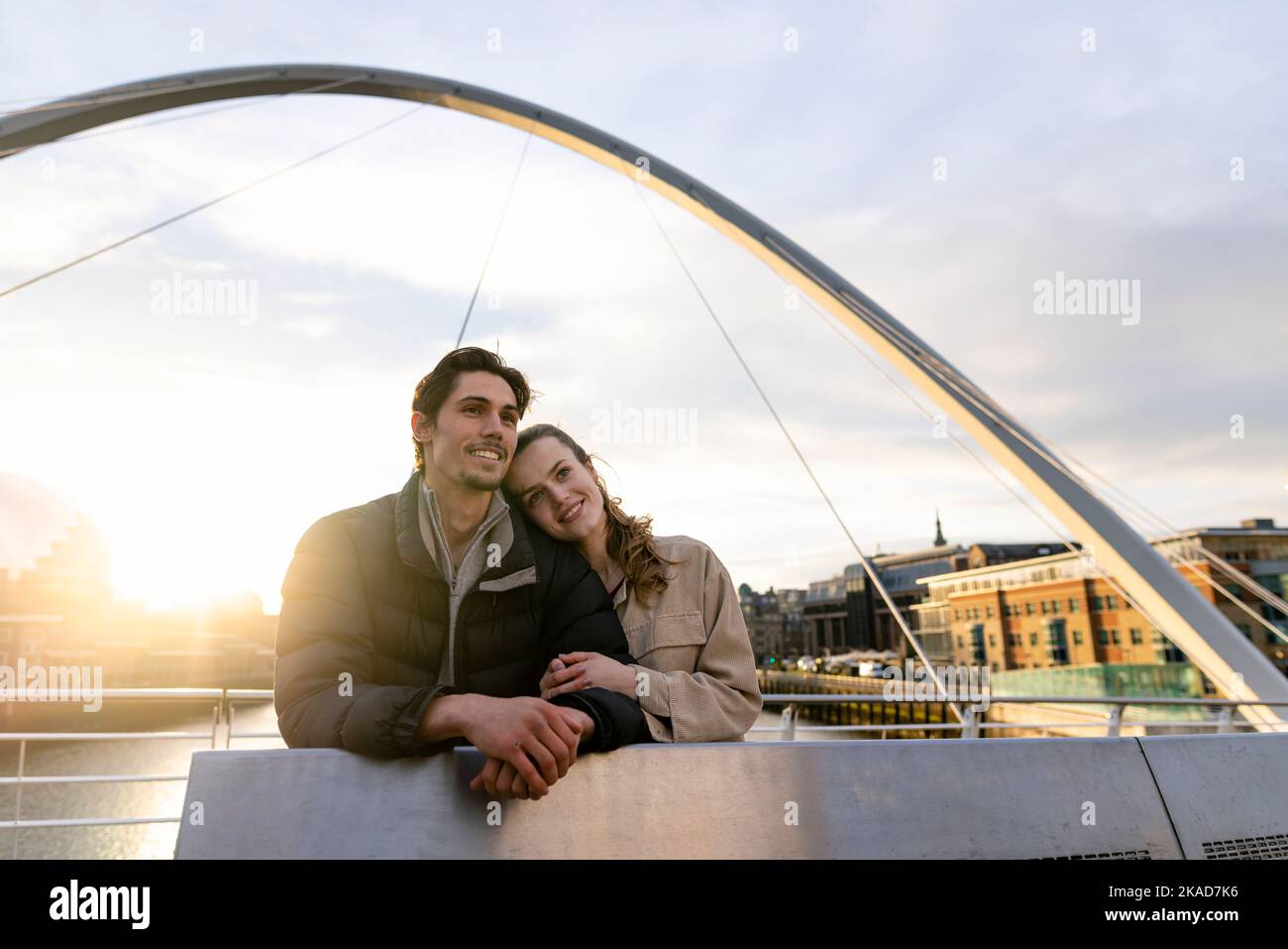 A wide-view shot of a young couple standing side by side looking at the view together on a bridge in the city, they are in love. Stock Photo