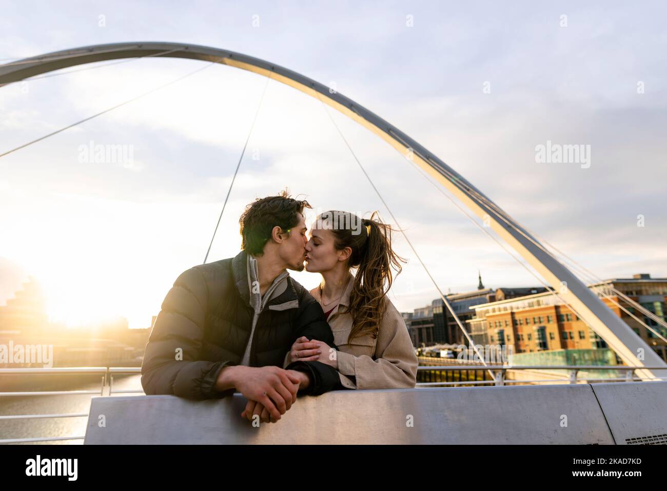 A wide-view shot of a young couple standing side by side sharing a kiss together on a bridge in the city, they are in love. Stock Photo