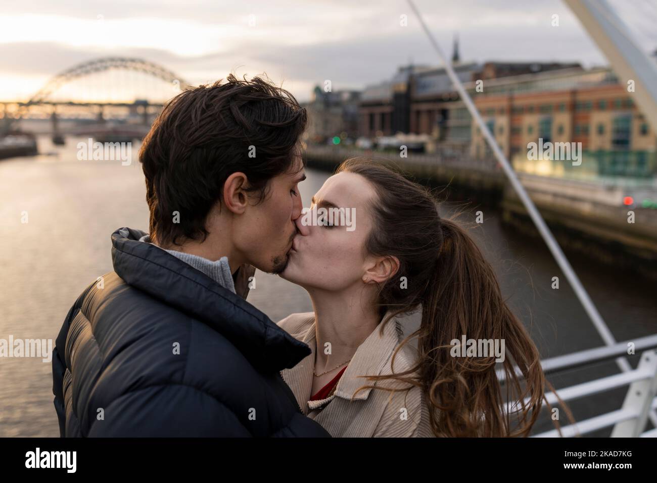 A close-up shot of a young couple standing side by side sharing a kiss together on a bridge in the city, they are in love. Stock Photo