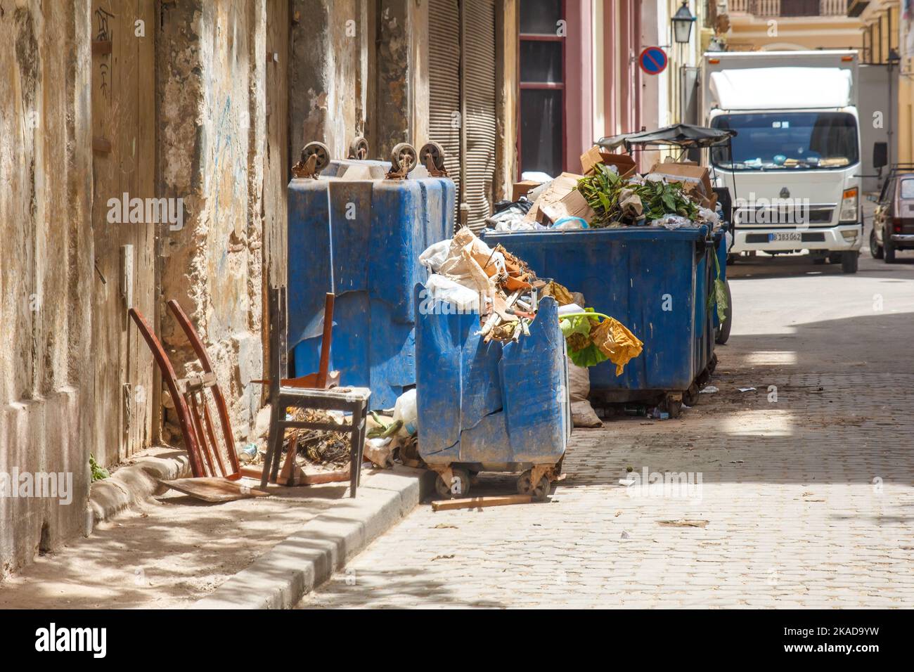 Overflowing urban garbage bins by run-down, weathered buildings. Some furniture is in the sidewalk. The street is made of cobblestone Stock Photo