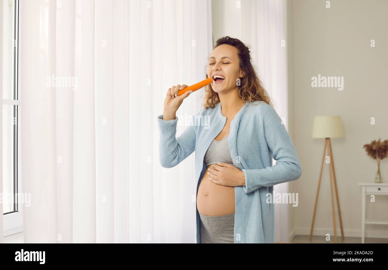 Happy smiling young pregnant woman standing by the window and eating a fresh carrot Stock Photo