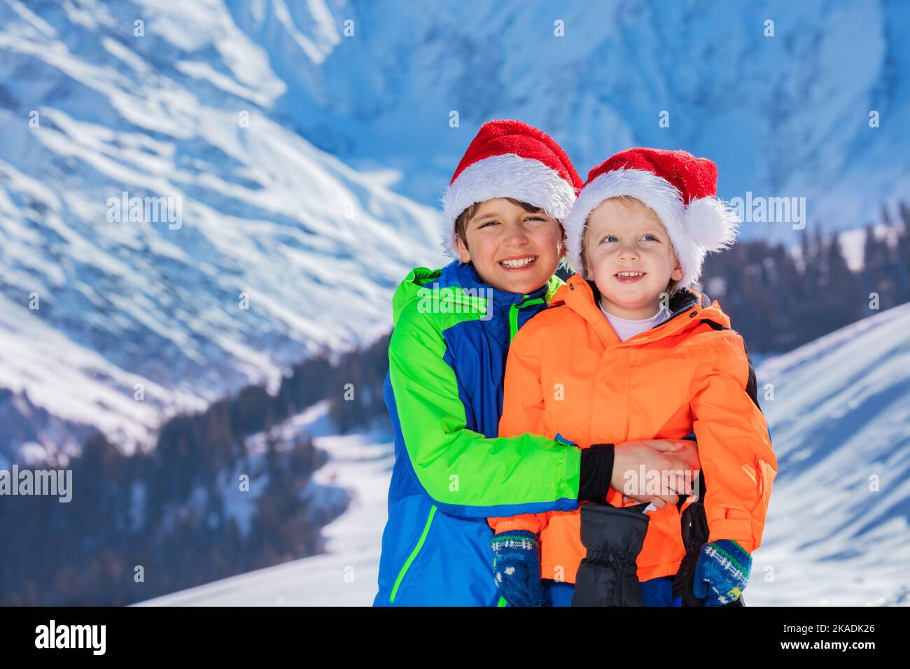Two happy boys in Santa Claus hats stand over snow mountains Stock Photo