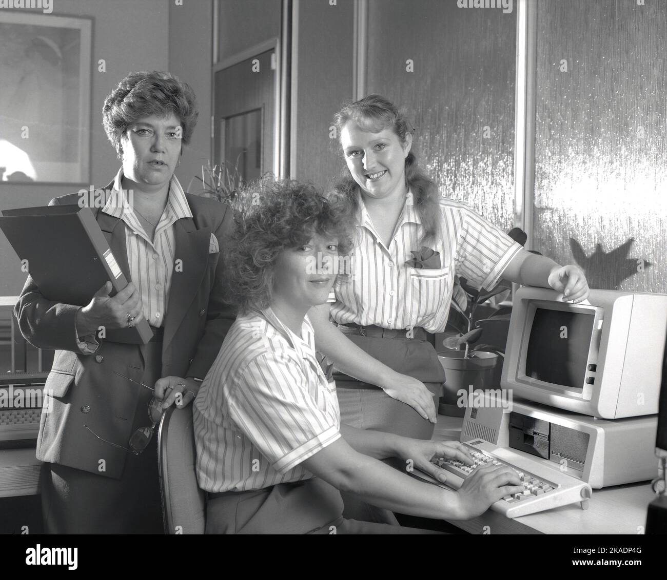 1989, historical, three office female staff by a computer of the era, England, UK. Manager in jacket behind, with folder, labeled 'House Rules'. One of the women is sitting at a keyboard infront of an IBM personal computer of the era, consisting of a separate display monitor and computer, with floppy disk capacity. Stock Photo