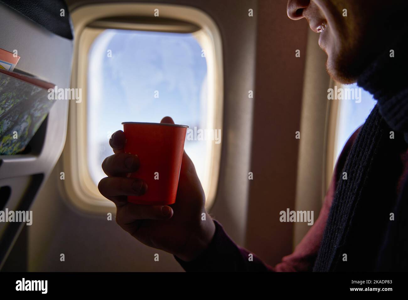 Smiling airline passenger with a paper cup of coffee looking ahead in airplane window. Man drinks tea onboard. Male holds beverage or water during Stock Photo