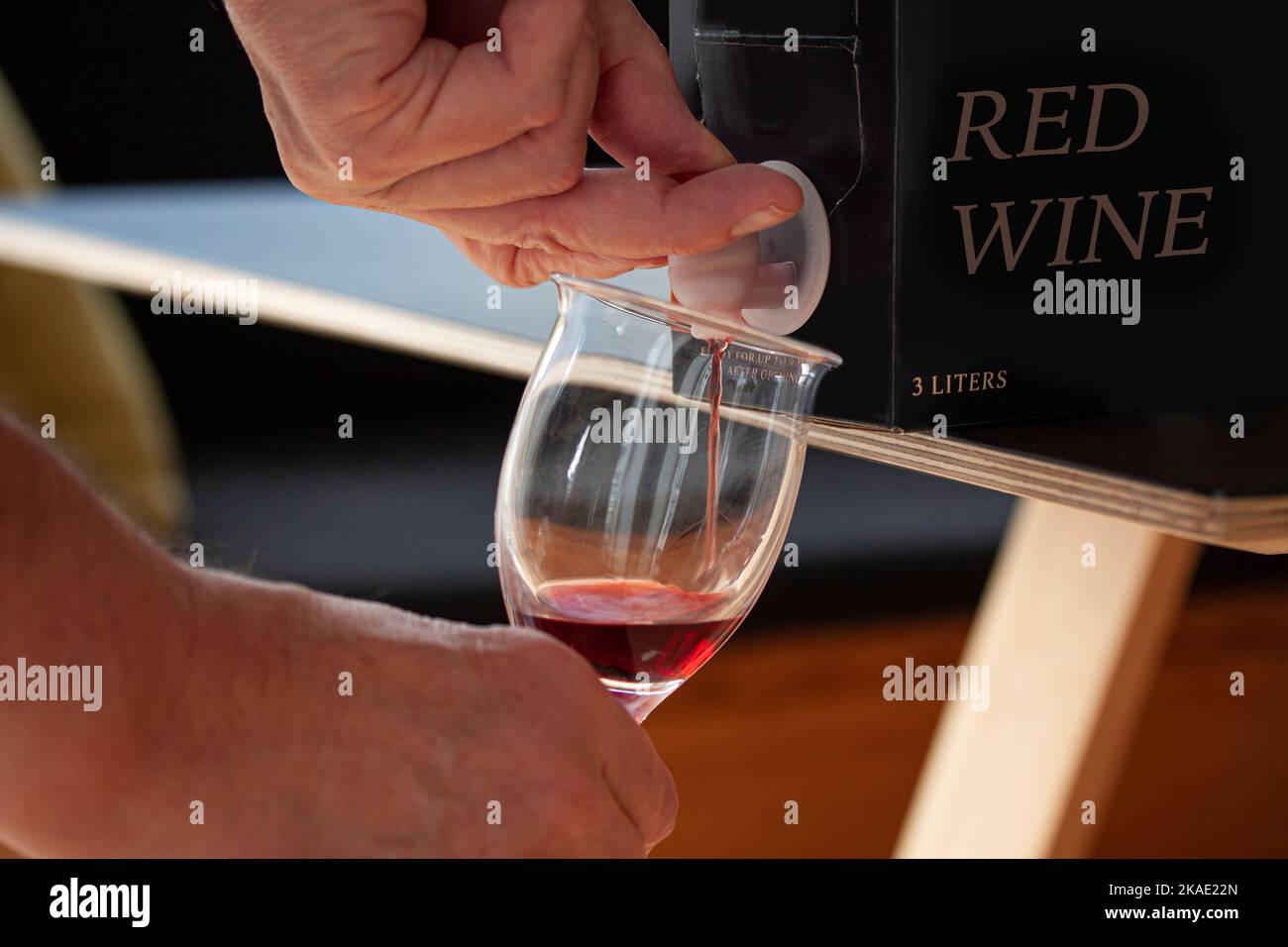 Hand pouring red wine into a glass from a BIB - cardboard bag in box with open tap standing on a table. Close up image. Fictitious brand. Stock Photo