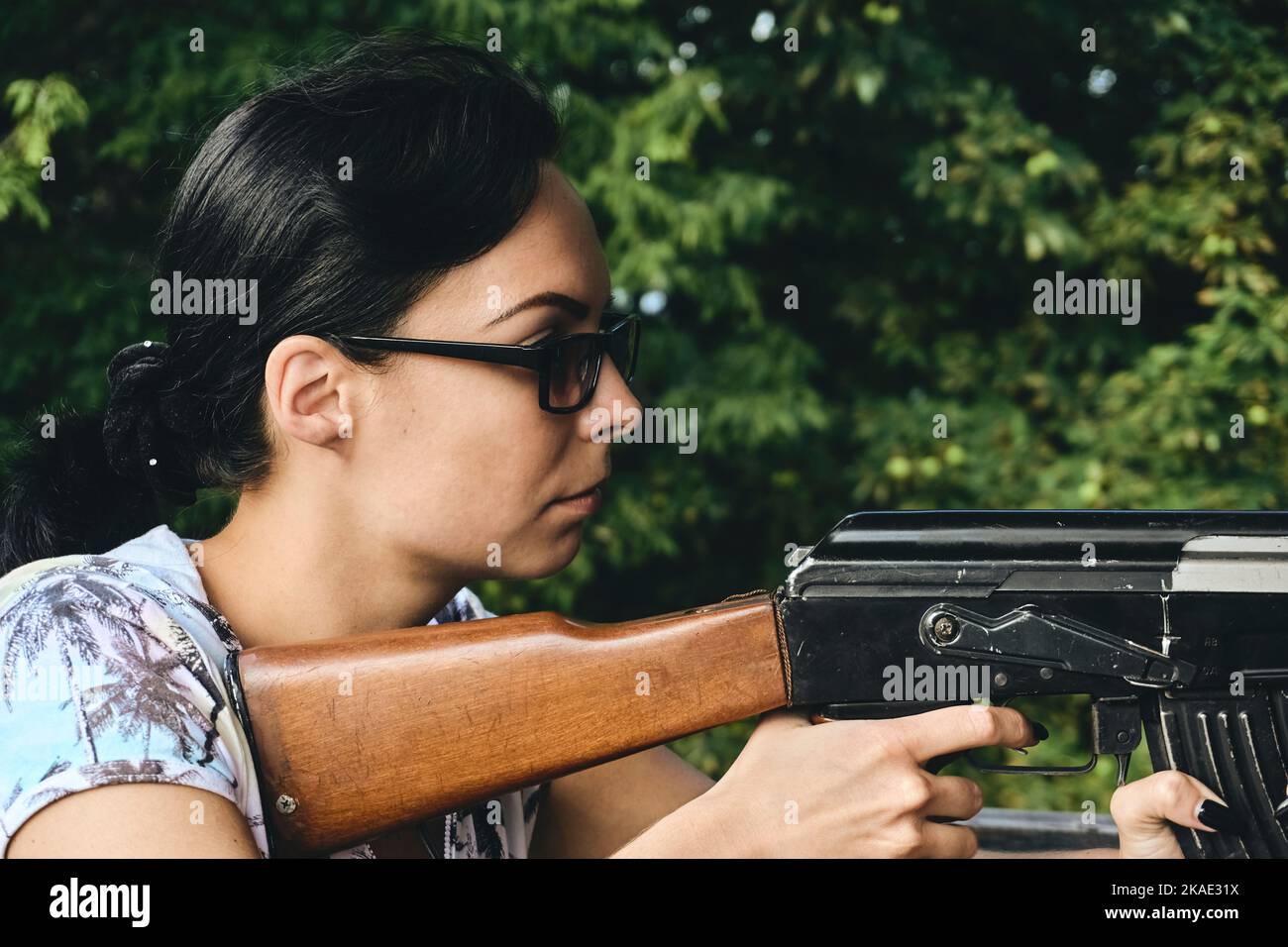 A young girl in dark sunglasses shoots from a machine gun and green trees Stock Photo