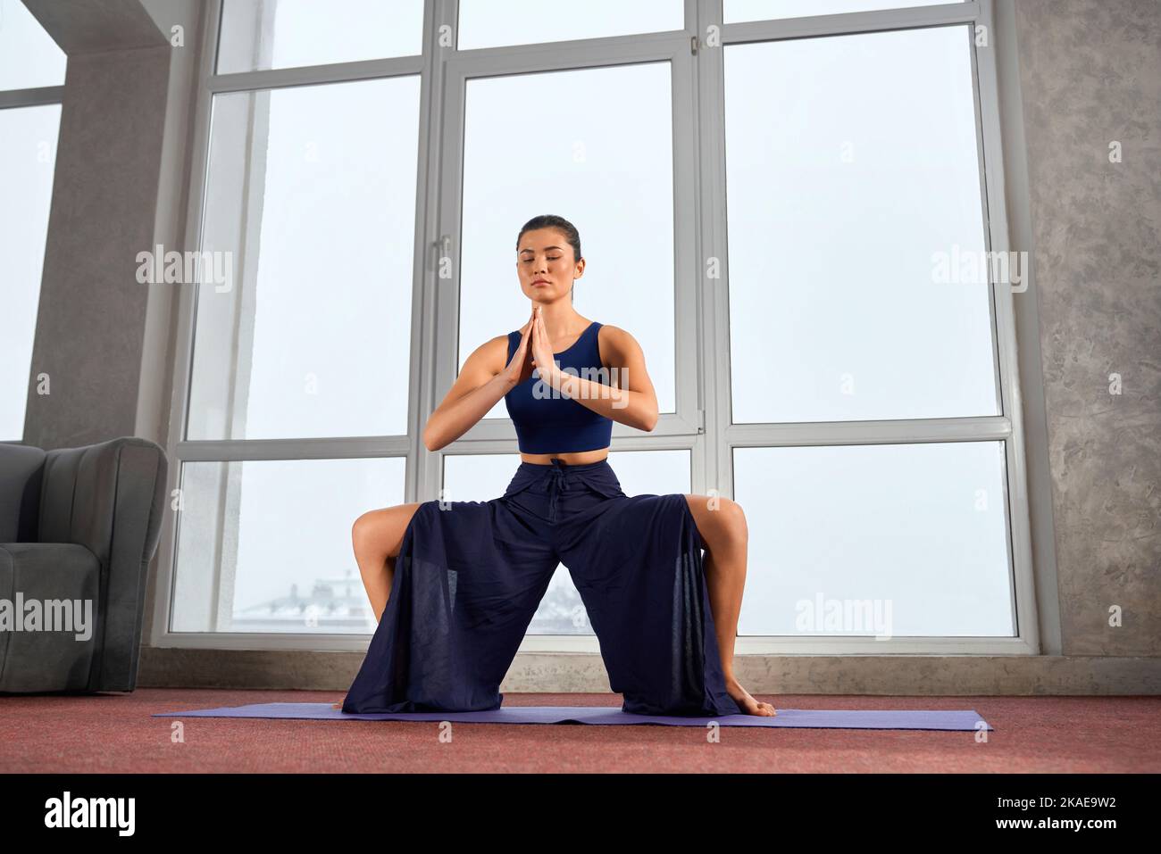 Side view of flexible girl sitting on bent legs, meditating indoors. Brunette female practicing yoga, with prayer hands and closed eyes, wearing palazzo pants. Concept of yoga. Stock Photo