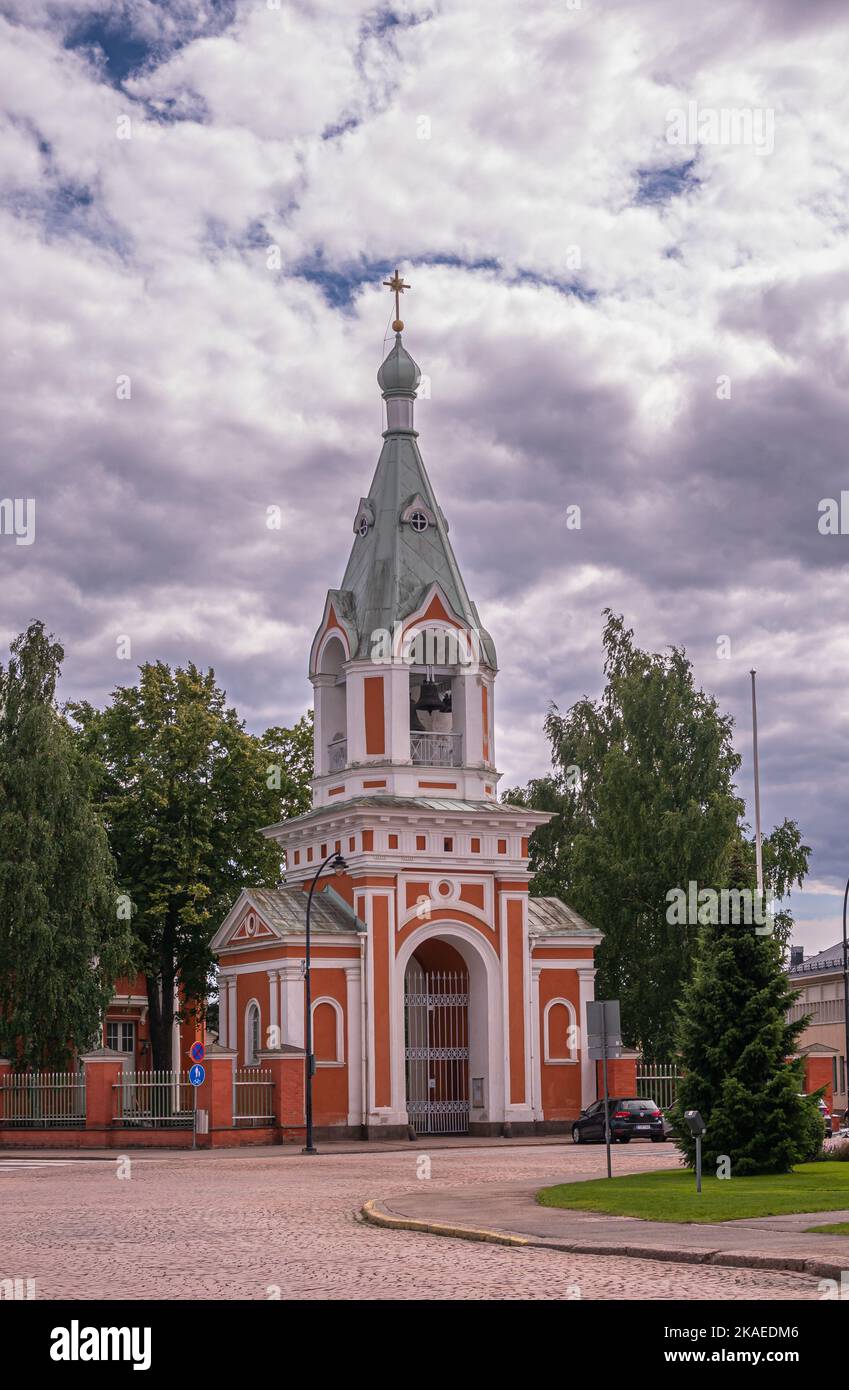 Finland, Hamina - July 18, 2022: Red and white Peter and Paul Church bell tower and entrance under heavy gray cloudscape with green foliage. Cars in s Stock Photo