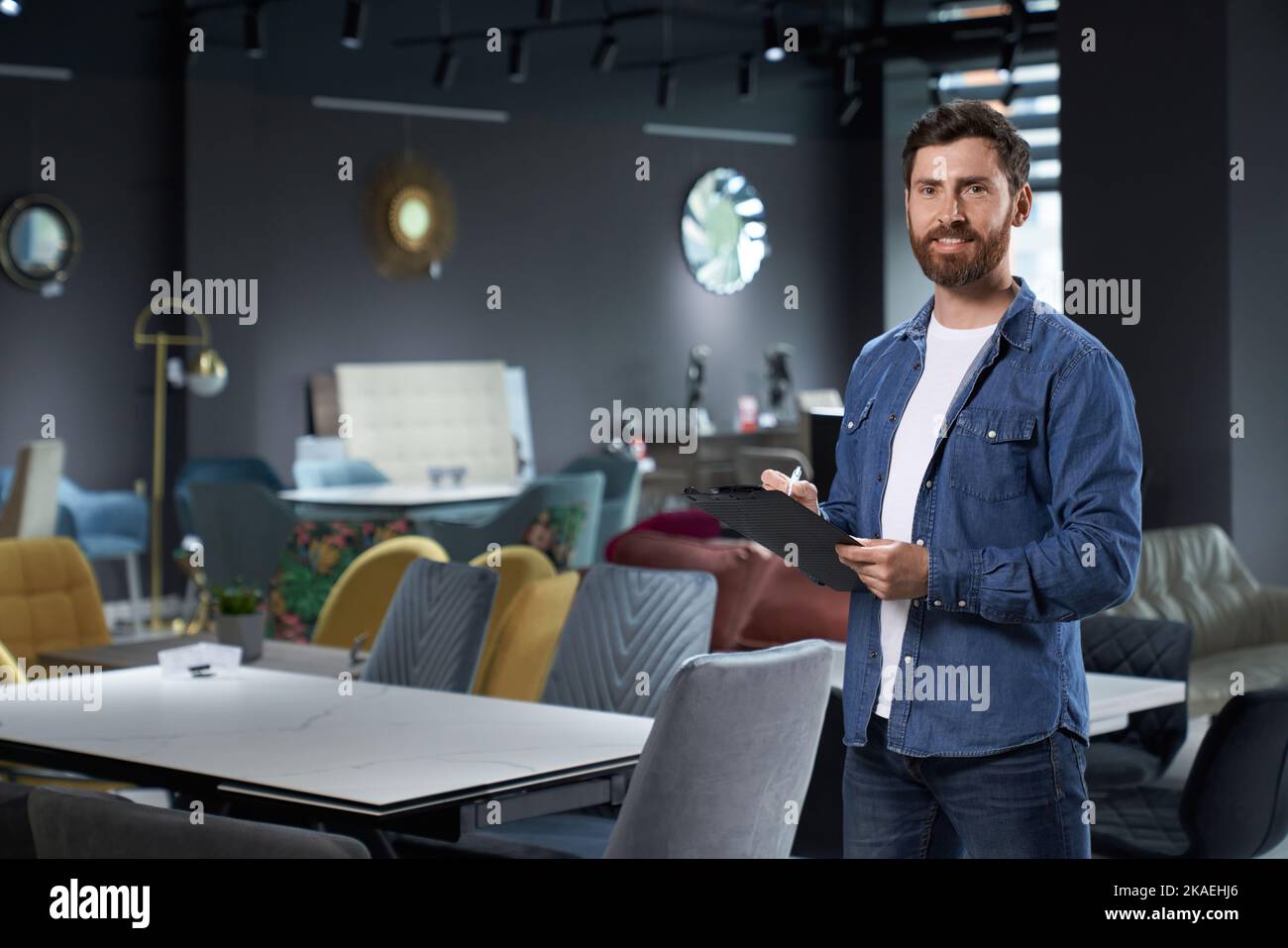 Smiling bearded salesman in denim standing with black tablet in new