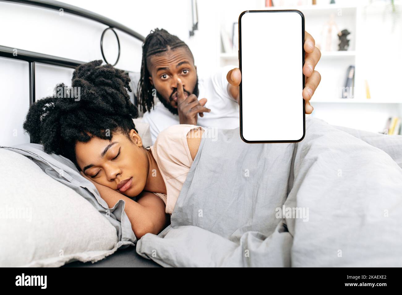 Mock-up concept. Happy african american spouses lie together in a cozy bed, wife sleeps and husband shows smartphone with blank white mockup screen, shows shhh gesture, prepares surprise for wife Stock Photo
