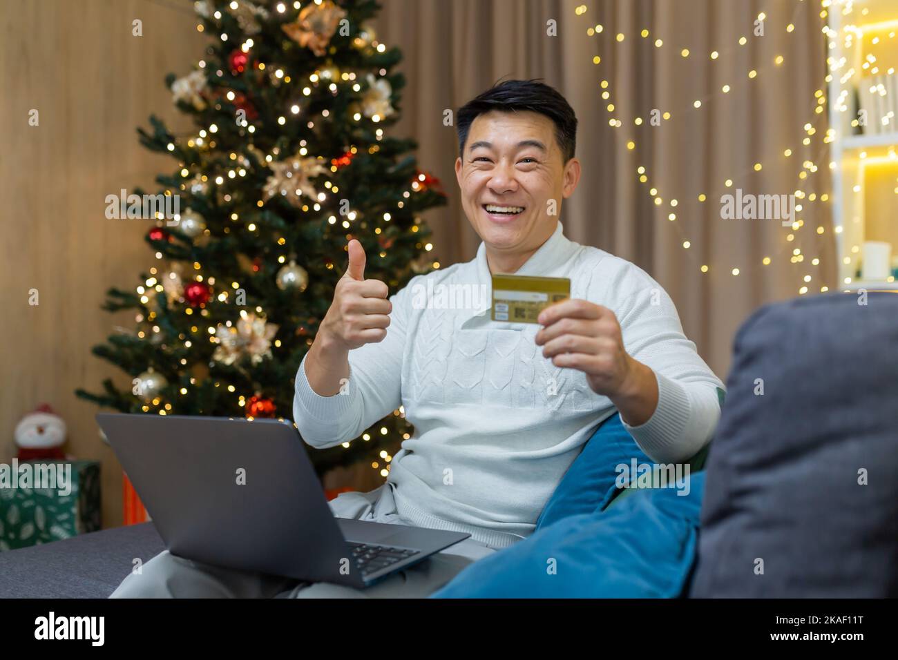 A young Asian man is sitting on the sofa near the Christmas tree at home. He is holding a laptop and a credit card. Shopping for Christmas gifts. He looks at the camera, smiles, points a super finger. Stock Photo