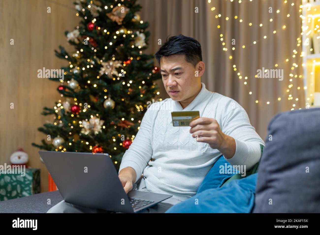 Confused and worried young handsome Asian man sitting at home and on sofa near Christmas tree. Holds laptop and credit card, can't buy gifts online, blocked card, ran out of money. Stock Photo