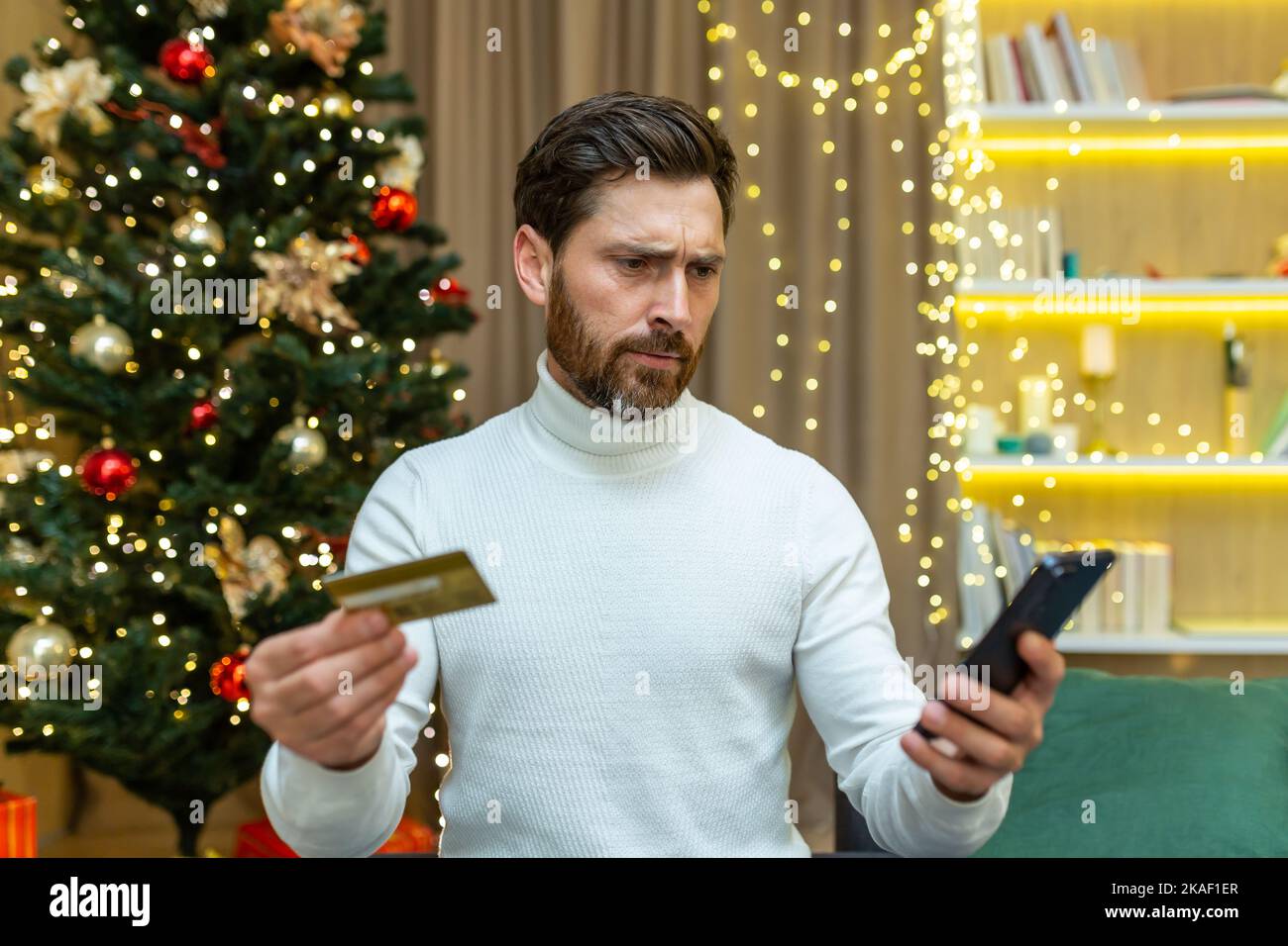 Confused and embarrassed man looks at the phone, holds a credit card in his hands. Sitting on the sofa near the Christmas tree, unable to make festive online purchases, he doesn't have enough money. Stock Photo