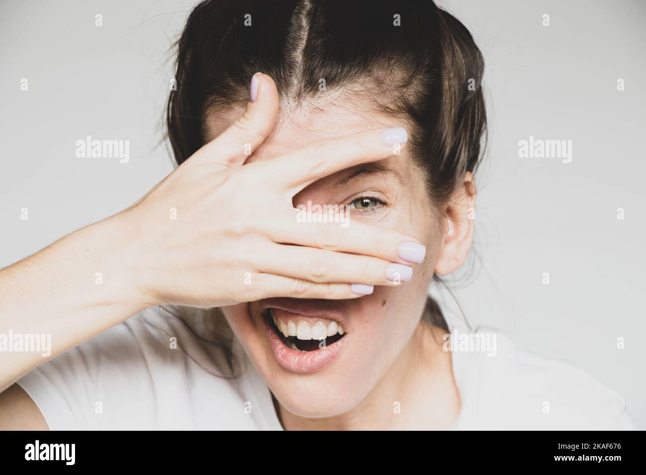 The girl closes one eye with her hand on a white background, looks through her hand, I see, I look Stock Photo