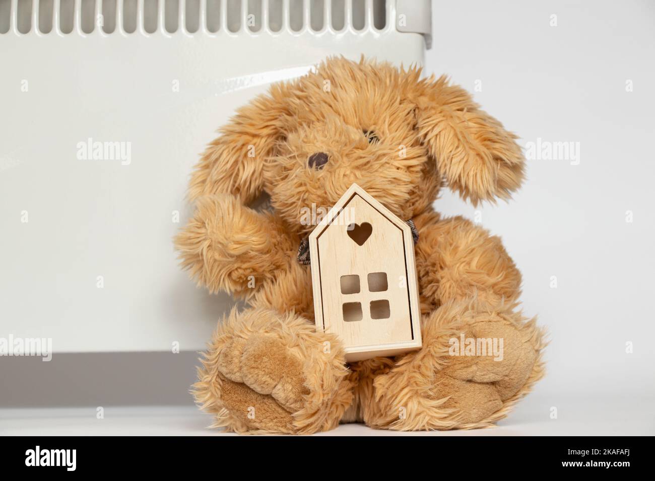 A plush hare and a small wooden house lie near the convector on a white background, a warm house and a heater, home heating appliances Stock Photo