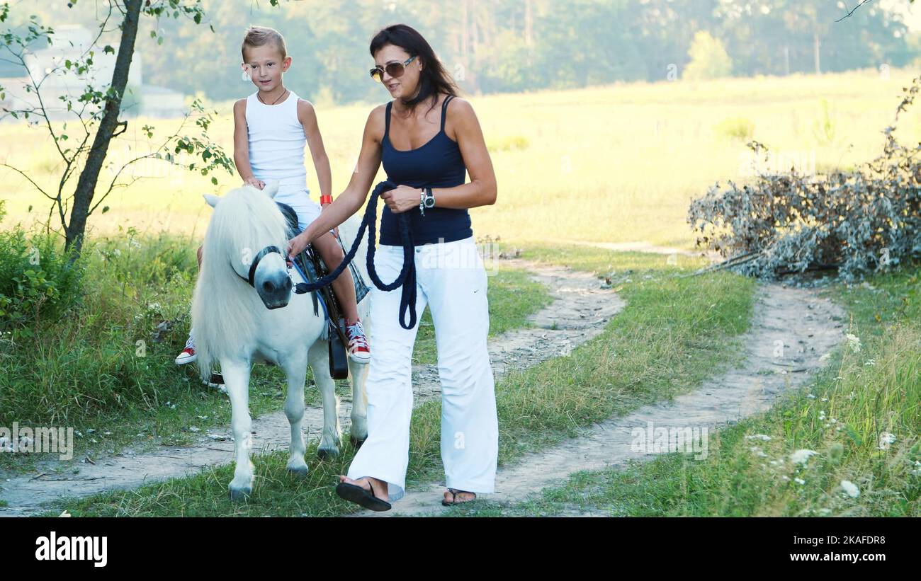 Mom and daughter are walking around the field, daughter is riding a pony, mother is holding a pony for a bridle. Cheerful, happy family vacation. Outdoors, in summer, near the forest. High quality photo Stock Photo