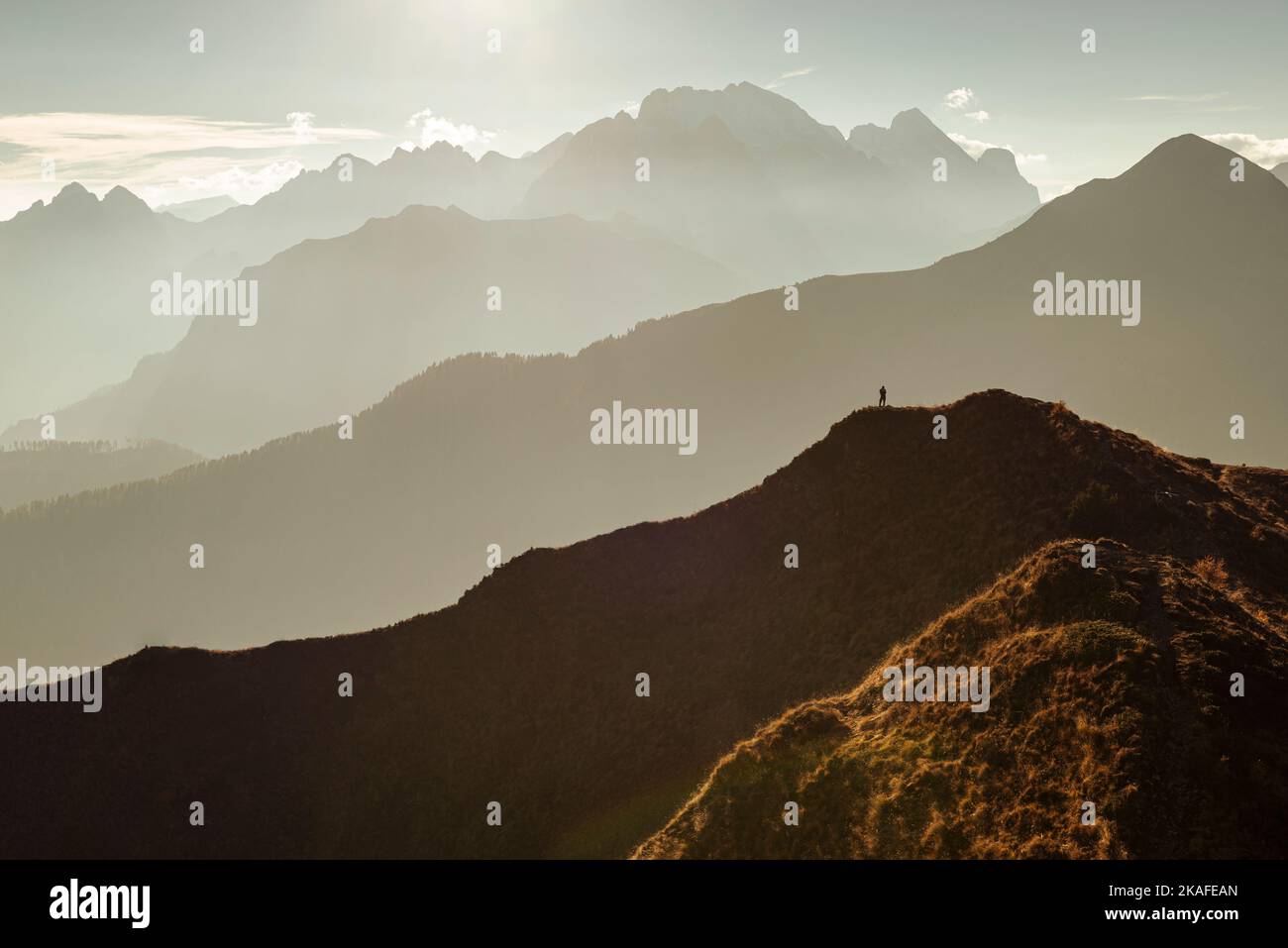 A man stands on a panoramic peak at Passo di Giau in the warm backlight in front of mountain ranges and the Mount Marmolada, Dolomites, Italy Stock Photo
