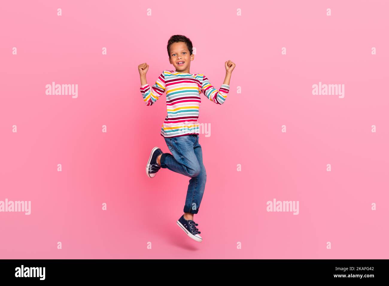 Full length photo of cheerful optimistic adorable boy dressed striped shirt jeans jumping hold fists up isolated on pink color background Stock Photo