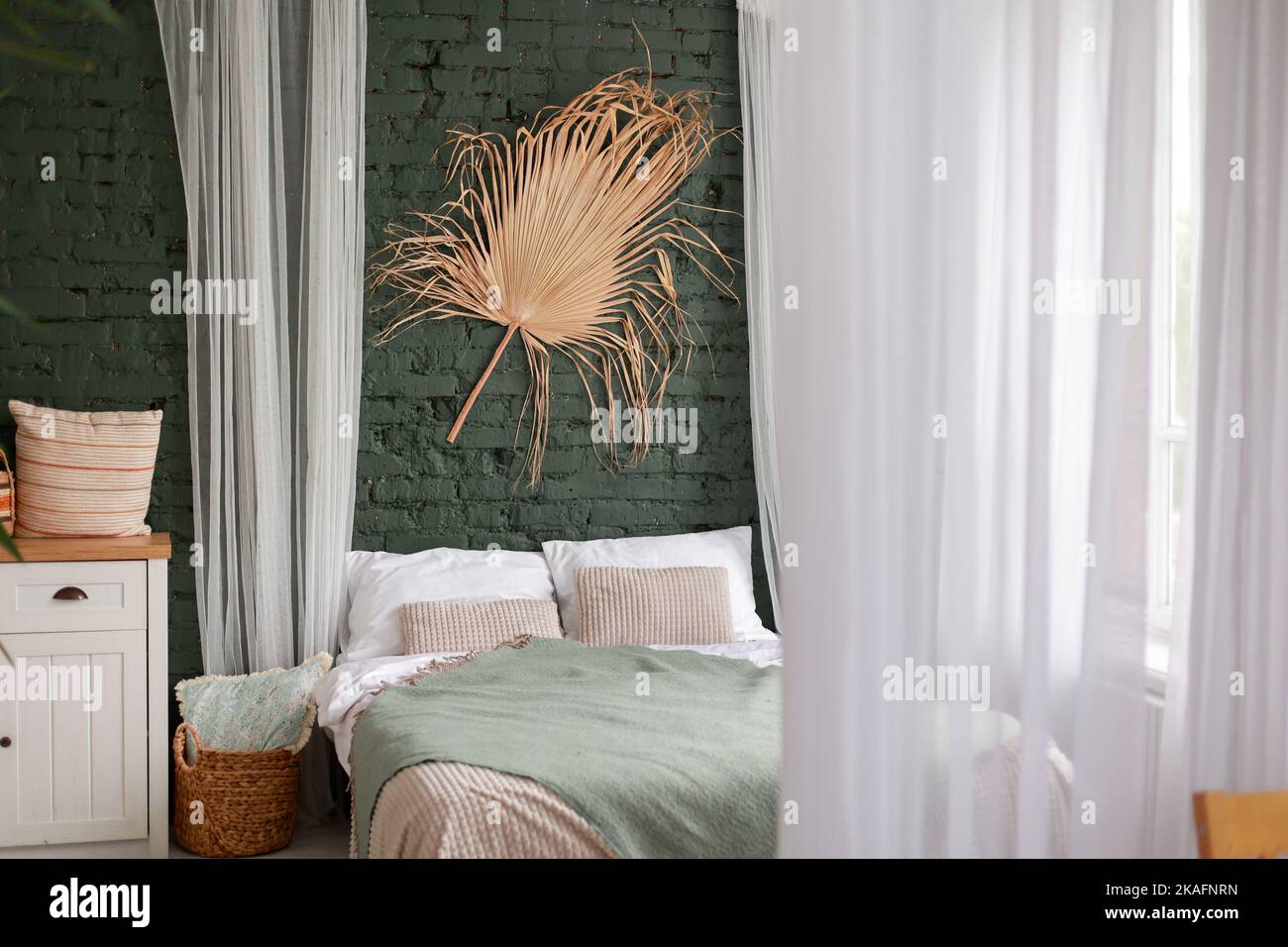 Modern boho interior of bedroom with green brick wall, bed, white wooden commode, curtains, houseplants, rattan basket, dray palm leaf and design pers Stock Photo