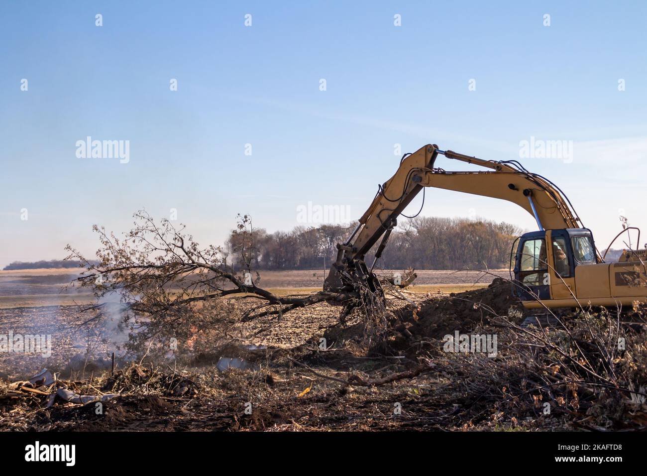 Close up view of a heavy equipment excavator moving trees and wooden debris into a fire pit near the area of a demolished old farm building Stock Photo