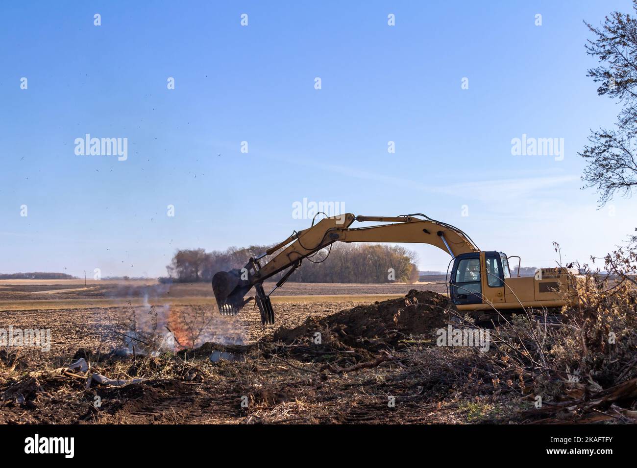 Close up view of a heavy equipment excavator moving trees and wooden debris into a fire pit near the area of a demolished old farm building Stock Photo