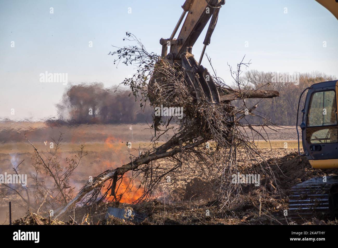 Close up view of a heavy equipment excavator moving trees and wooden debris into a fire pit near the area of a demolished old farm building Stock Photo