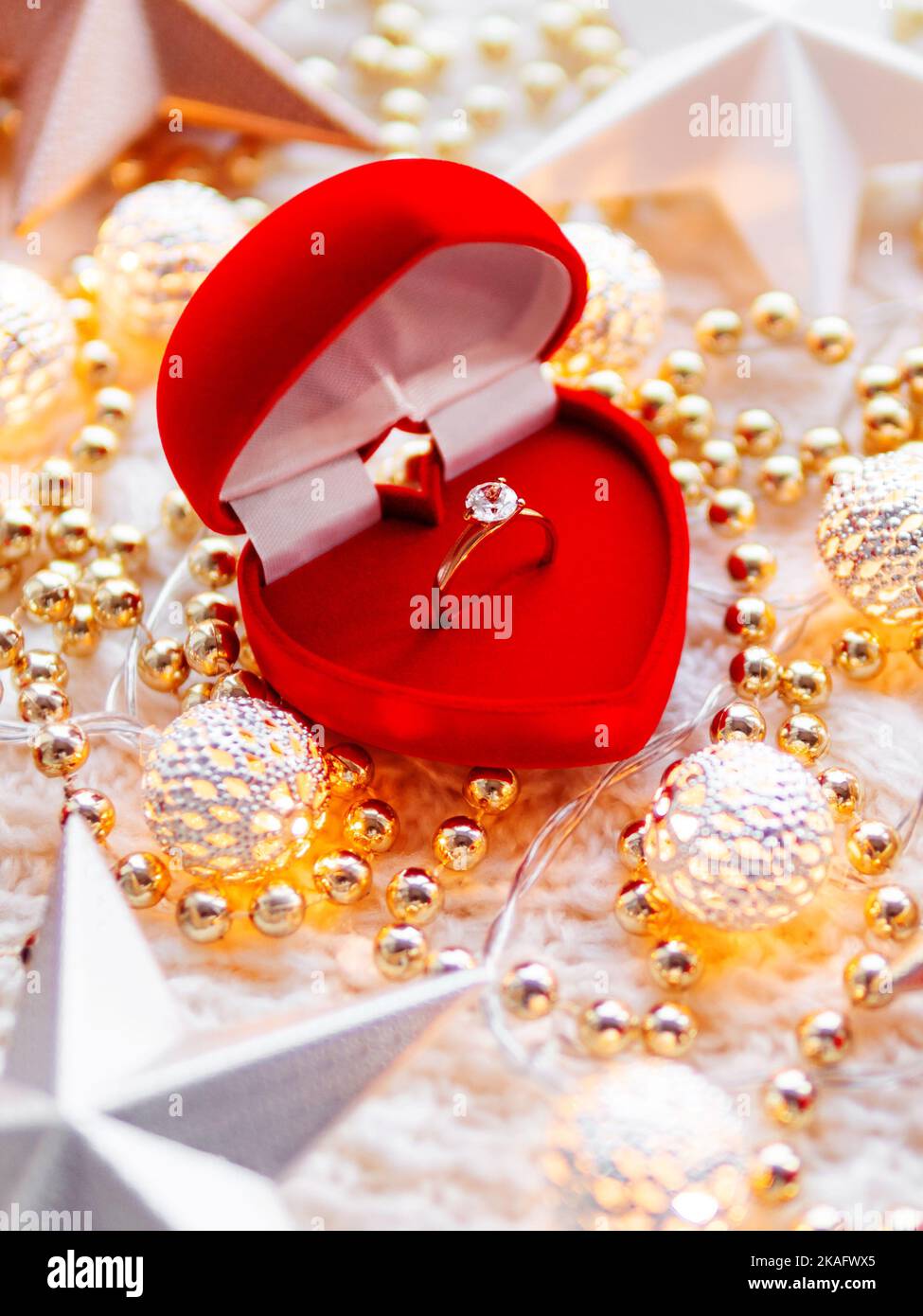 Christmas and New Year star decorations on white knitted background. Red heart gift box with engagement golden ring, metal light bulbs Stock Photo