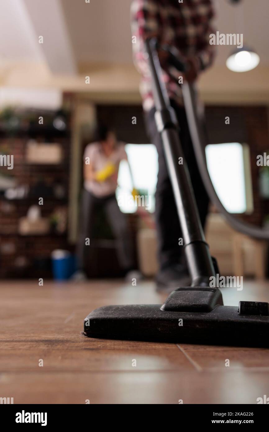 Man vacuuming apartment floor while woman in background dusts shelves. Modern couple doing household chores, finishing spring cleaning and disinfecting spaces, out of focus image. Stock Photo