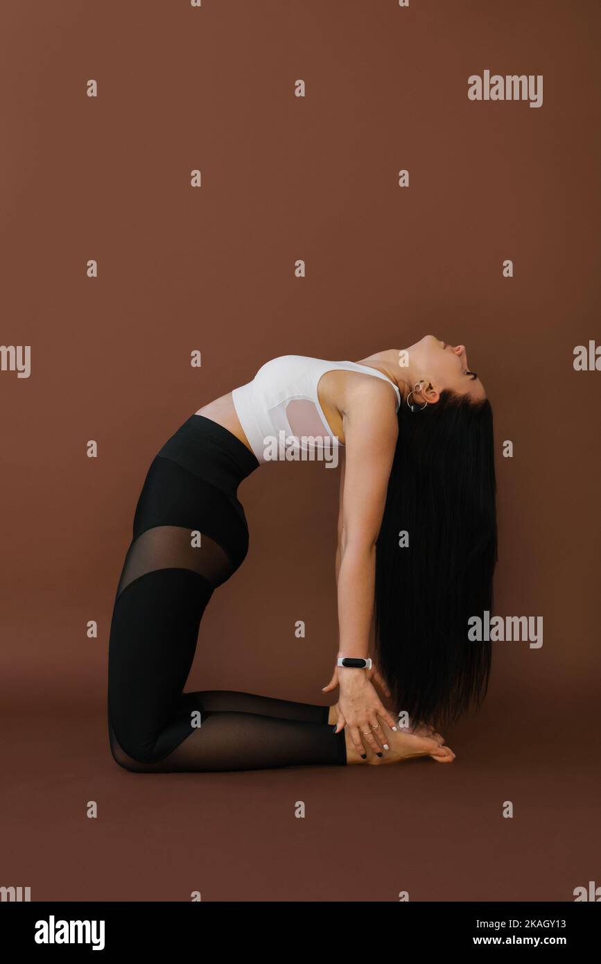 Young Caucasian woman doing yoga, stretching in the exercise is arranged, camel pose, workout, sportswear, white top and black trousers, indoor full-l Stock Photo