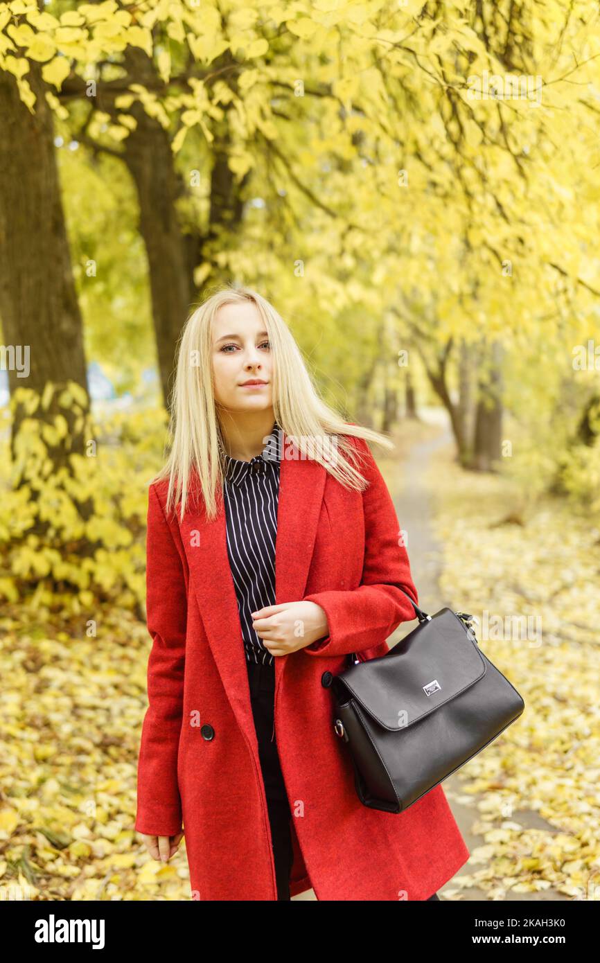 A young blonde woman walks around the autumn city in a red coat. The concept of urban style and lifestyle. Stock Photo