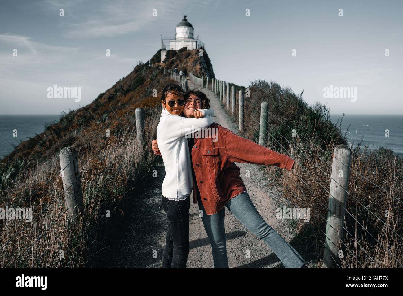 couple of brunette caucasian girls disheveled by the wind embracing happily having fun on the path that leads to the impressive lighthouse, nugget Stock Photo