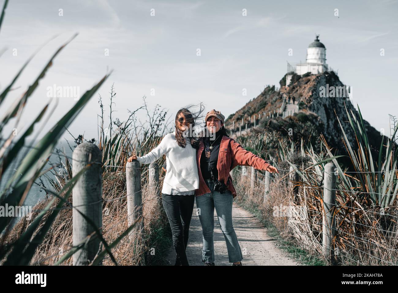 two caucasian girls in red jacket white sweater black and blue pants happy looking at camera standing on path leading to impressive lighthouse, nugget Stock Photo