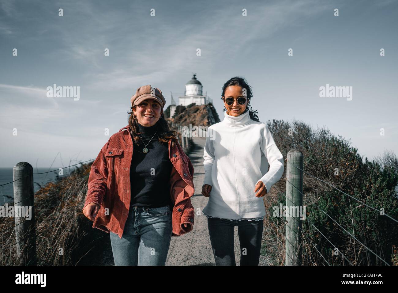 two young caucasian women running towards camera smiling and happy in red jacket beanie and white pullover with stunning lighthouse behind them Stock Photo