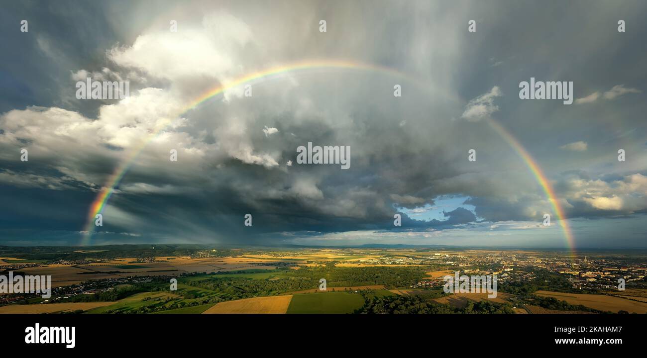 A rainbow over a farmland, the remnants of a storm, a dramatic sky, ripening cornfields, a town and hills in the background. Aerial panoramic view. Stock Photo