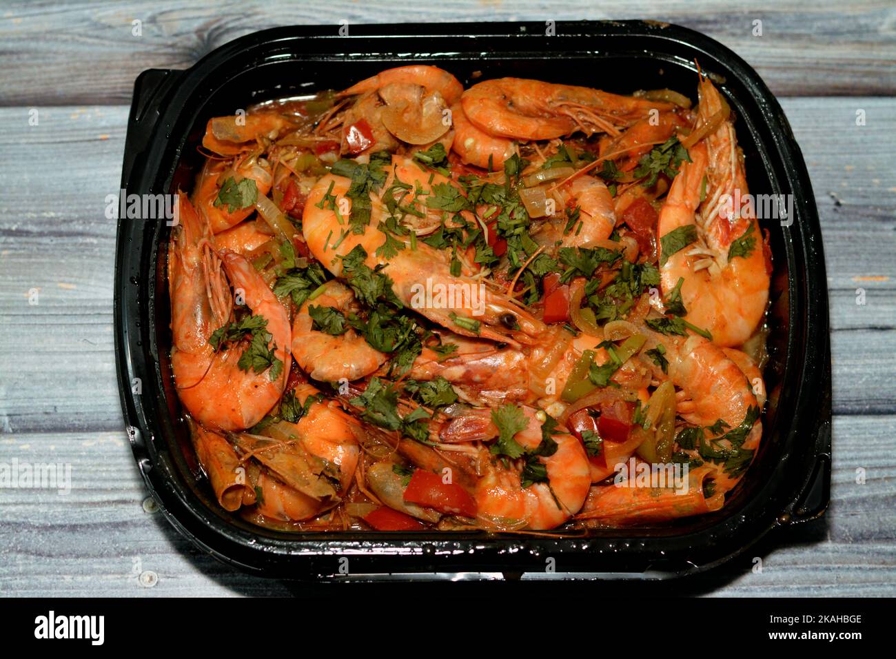 Boiled shrimps mixed garlic, lemon, parsley, pepper, onion and tomato served in disposable black plate isolated on wooden background, selective focus Stock Photo