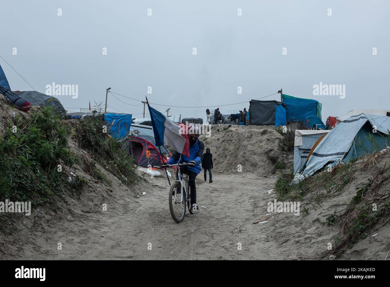 A migrant drives trough the Calais Jungle on a bike with a french flag. After registration, the migrants are distributed on buses. The refugee camp on the coast to the English Channel is to be cleared today. The approximately 8,000 refugees are distributed after the registration by busses to various reception centers in France, on October 24, 2016. (Photo by Markus Heine/NurPhoto) *** Please Use Credit from Credit Field *** Stock Photo