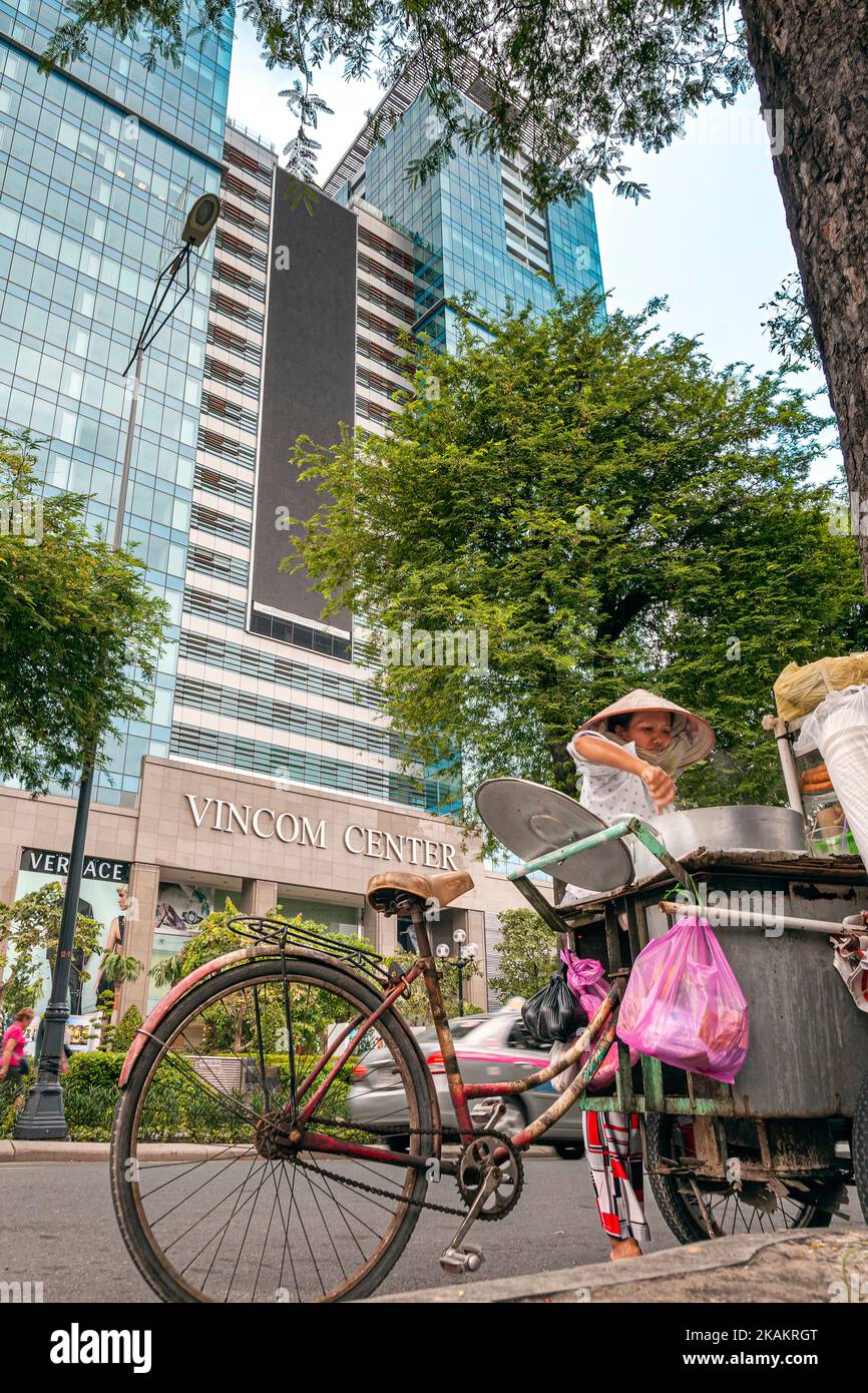 Contrast between traditional old Vietnamese lady wearing bamboo hat selling food on the street, and modern Vincom Centre Shopping Mall across road, Ho Stock Photo