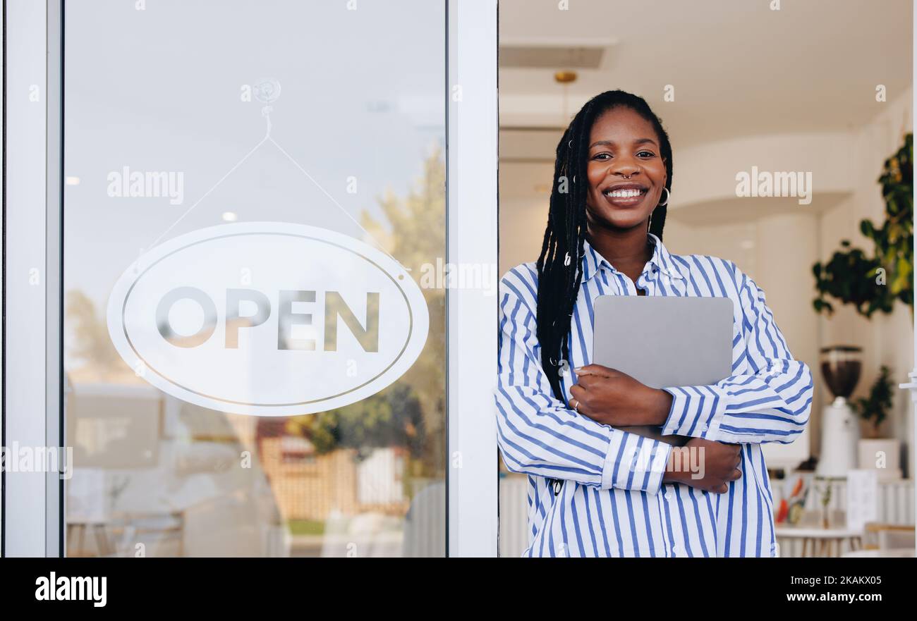 Ambitious young coffee shop owner standing next to an open sign at the entrance of her new restaurant. Female small business owner smiling at the came Stock Photo