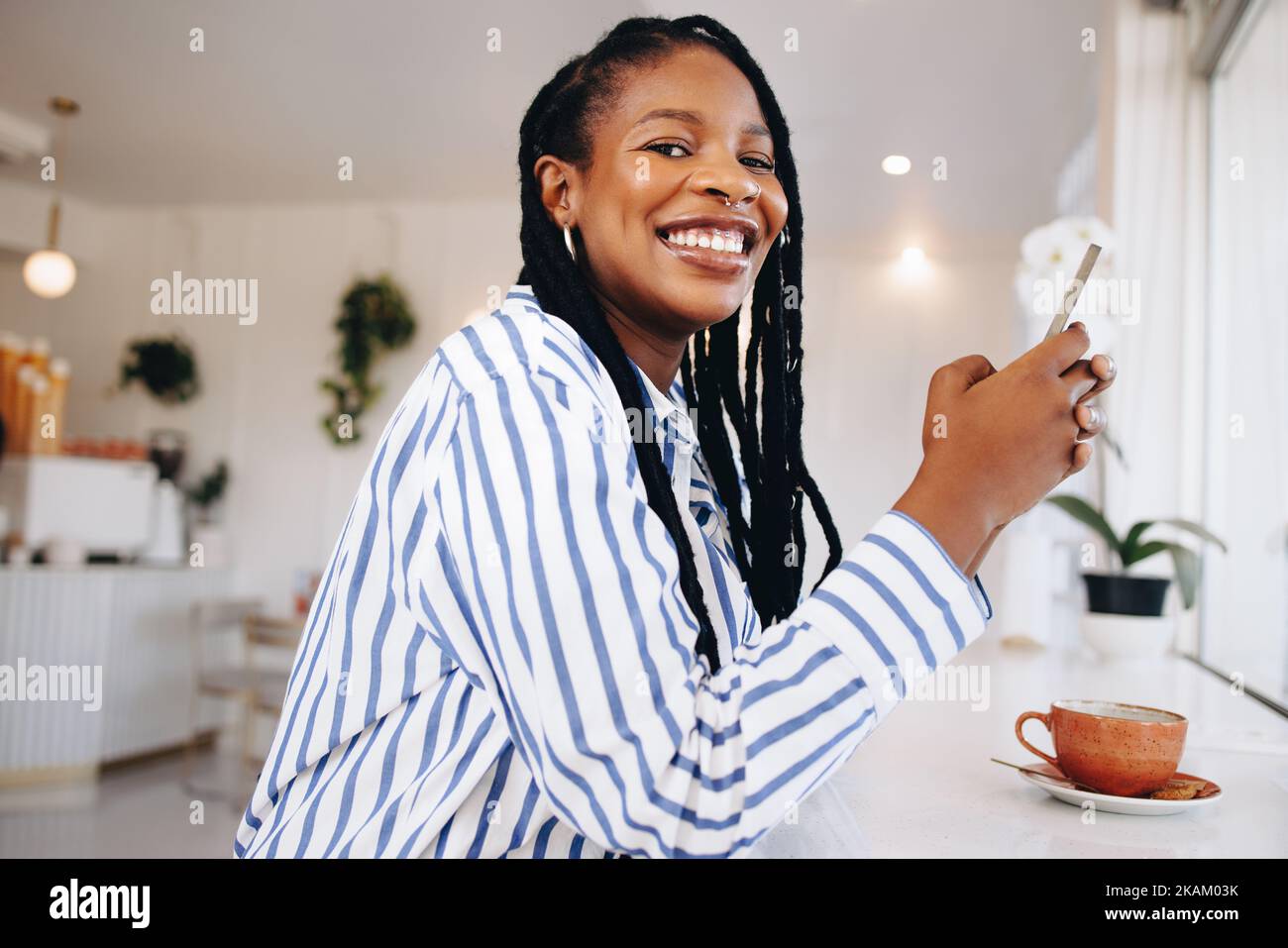 Happy young businesswoman smiling at the camera while holding a smartphone in a coffee shop. Cheerful young black businesswoman taking a coffee break Stock Photo
