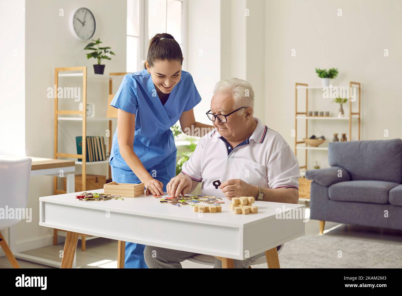 Smiling young female social worker and old man playing with jigsaw puzzle in nursing home. Stock Photo