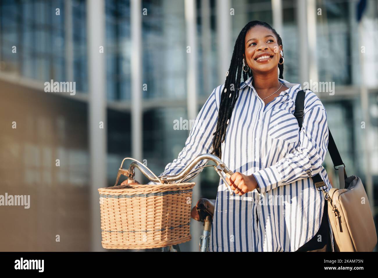 Smiling business woman looking away pensively while walking alongside her bicycle in the city. Cheerful young business woman commuting to her office i Stock Photo