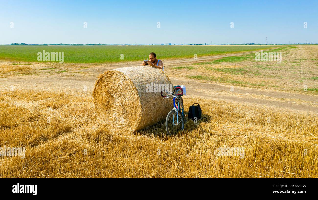 Above view, artist is holding remote, wireless, console to control drone motion with joystick leaned on round bale of straw at agricultural field. Stock Photo