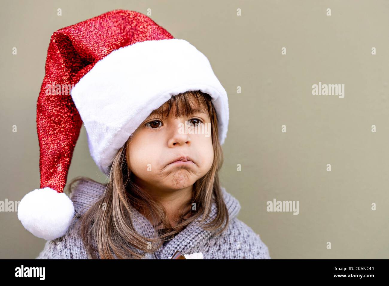 Cute little girl wearing Santa Claus hat outdoors in christmas time - Little girl sulking for Xmas gift - Funny face, Grinch style, grimace, childhood Stock Photo