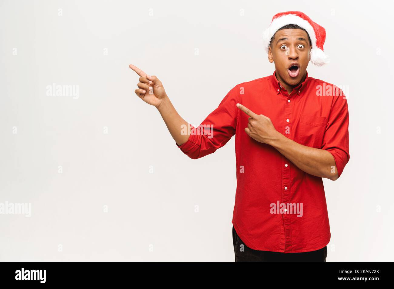 Excited multiracial young man wearing red santa hat points fingers aside at empty copy space, presenting novelty, advertising product. Surprised shocked guy promoting new year deal, holiday sale Stock Photo